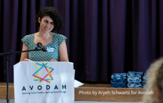 Ellen Oshinsky, a young woman with dark curly hair, is standing and smiling behind a podium with the Avodah logo and the words Avodah, Sparking Jewish Leaders, Igniting Social Change. Photo by Aryeh Schwartz for Avodah.